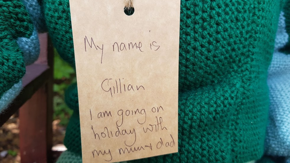 Label on knitted character, reads: My name is Gillian, I am on holiday with my mum and dad