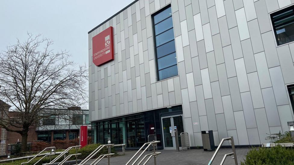 The front entrance of a large grey university building, with a red sign on it that says "Staffordshire University".