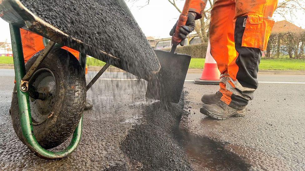 A pothole being repaired by a workman wearing hi vis orange overalls. He is pouring Tarmac out of a wheelbarrow into a pothole in the middle of a road.