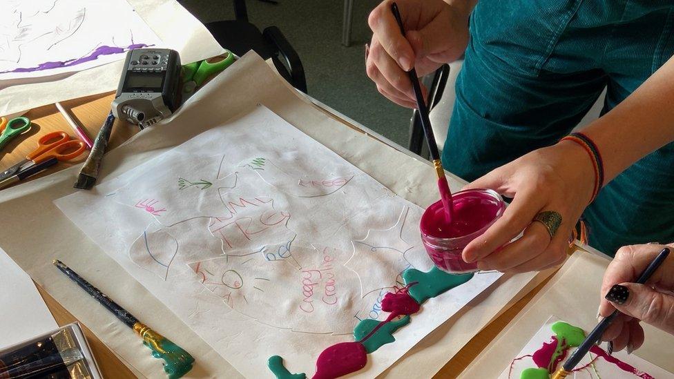 A piece of artwork with a person's hands in shot, dipping a paintbrush into pink paint