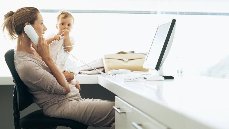 Posed photo of a woman on the phone at an office desk and holding a baby.