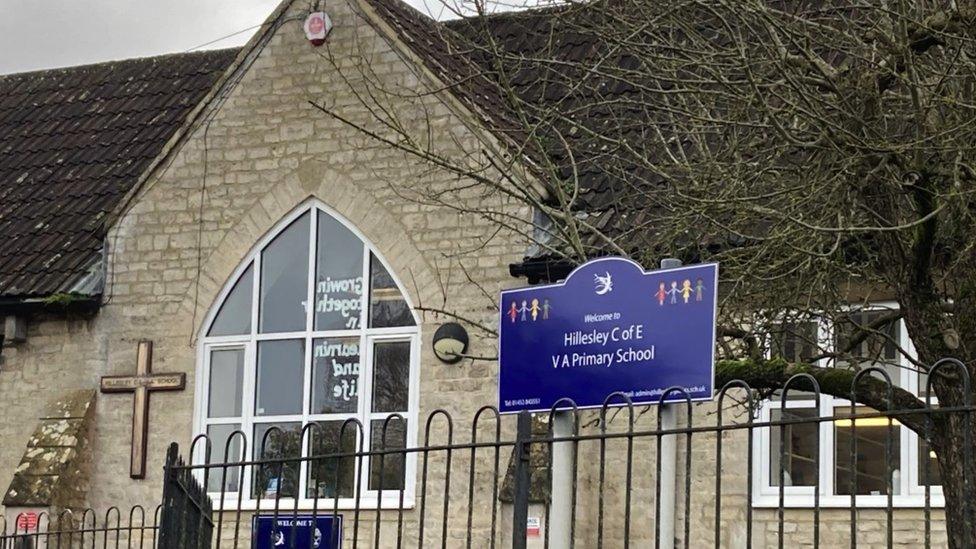 Exterior of Hillesley Primary School. It is built with Cotswold stone and there is a large window. A crucifix cross is on the wall next to the window. A blue sign reading "Welcome to Hillesley C of E V A Primary School" is in front of the building.