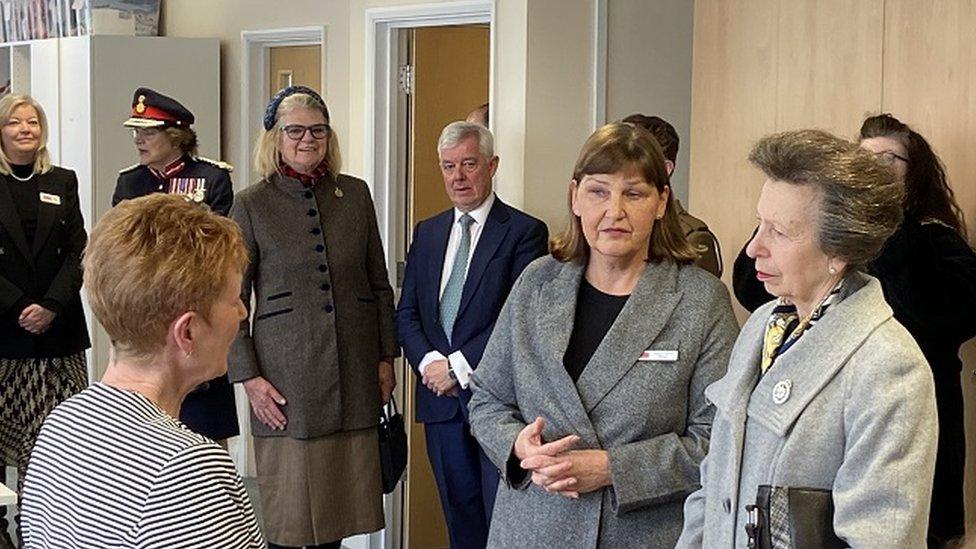 The Princess Royal surrounded by dignitaries listening to a volunteer talking through a garment