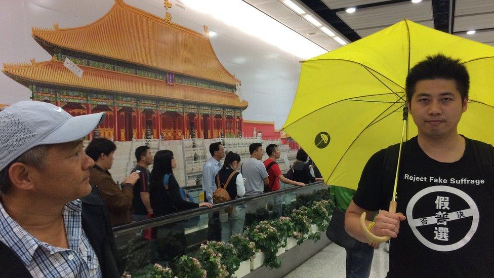 Billboard-sized image of the Forbidden City with passers-by and a man with a yellow umbrella and a T-shirt reading "reject fake suffrage", in front.