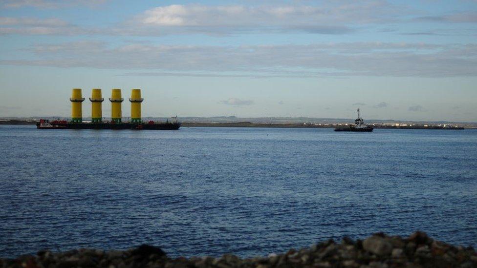 Four monopile structures used as the foundation of wind turbines are transported on a barge towed by a tug boat out of the mouth of the River Tees in Redcar in 2020