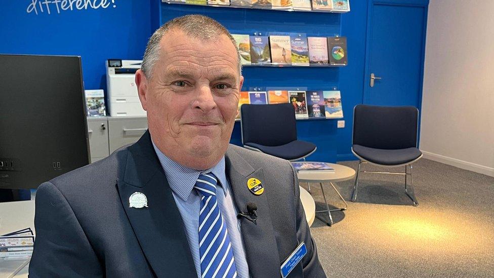 A portrait of Trevor Ridler, travel agent manager. He is smiling at the camera, sitting in his agency. Travel magazines are seen in the background.