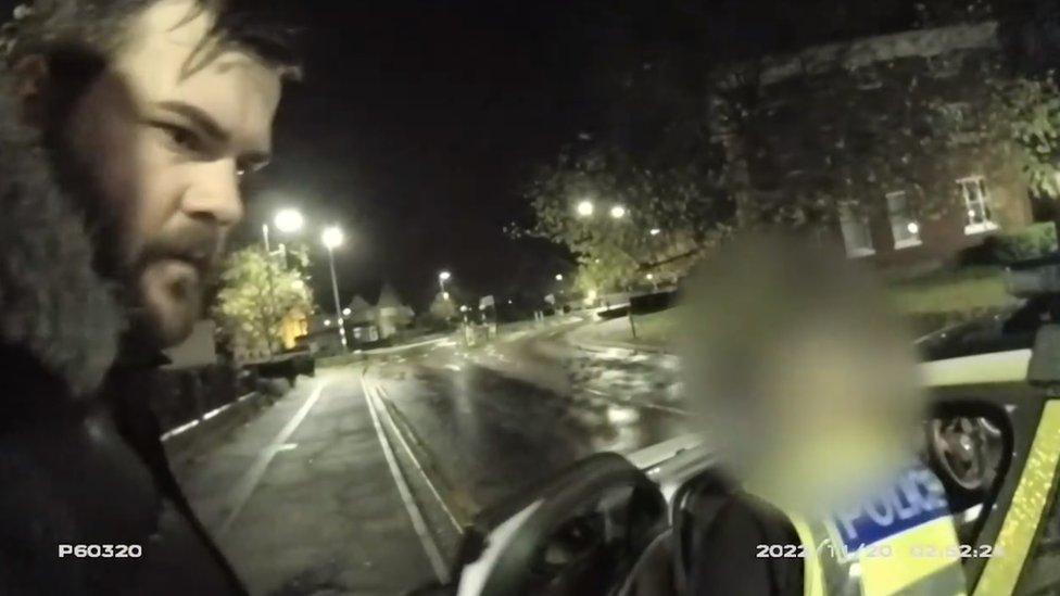 Man with dark beard wearing a coat with a furry hood is scene near a police car and an officer