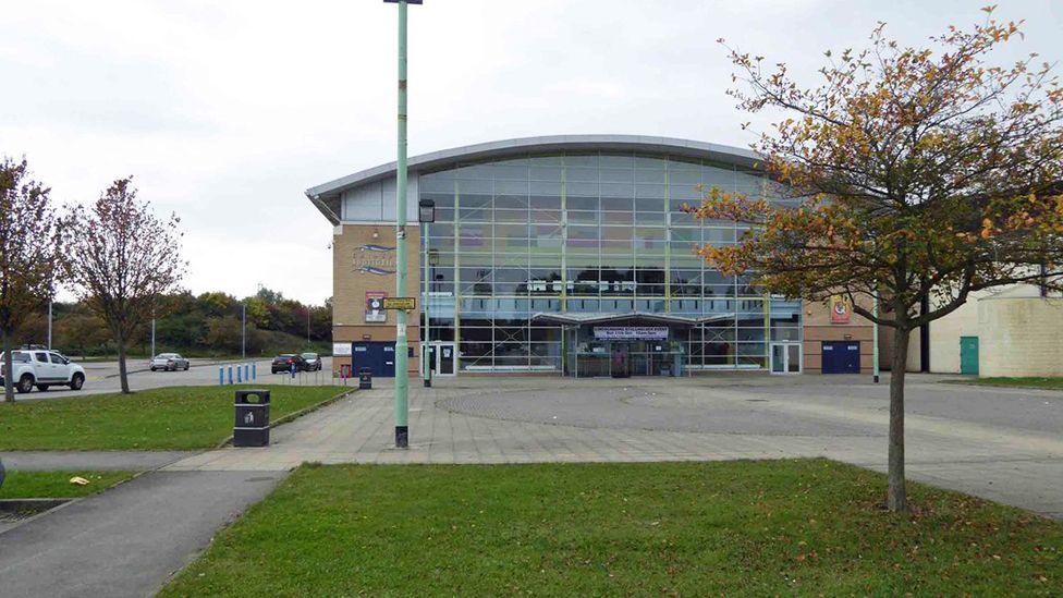 Outside of Grimsby Auditorium which has a glass front with brick on the side and a curved roof. There is a paved square in front of the building and a road running on the side
