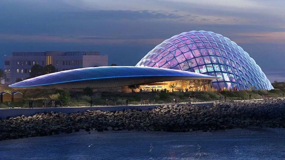 The curved roof of the proposed Eden Project Morecambe is seen in front of a glass domed, lattice framed building at the edge of a beach in an artist's drawing. To the left is a large roof over garden areas that is shaped like a long shell