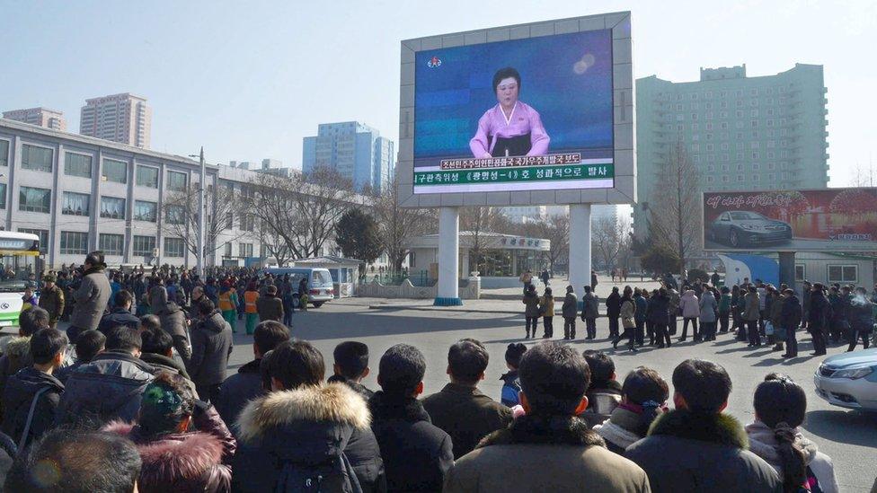 North Koreans watch a huge screen broadcasting an official announcement by a TV news presenter