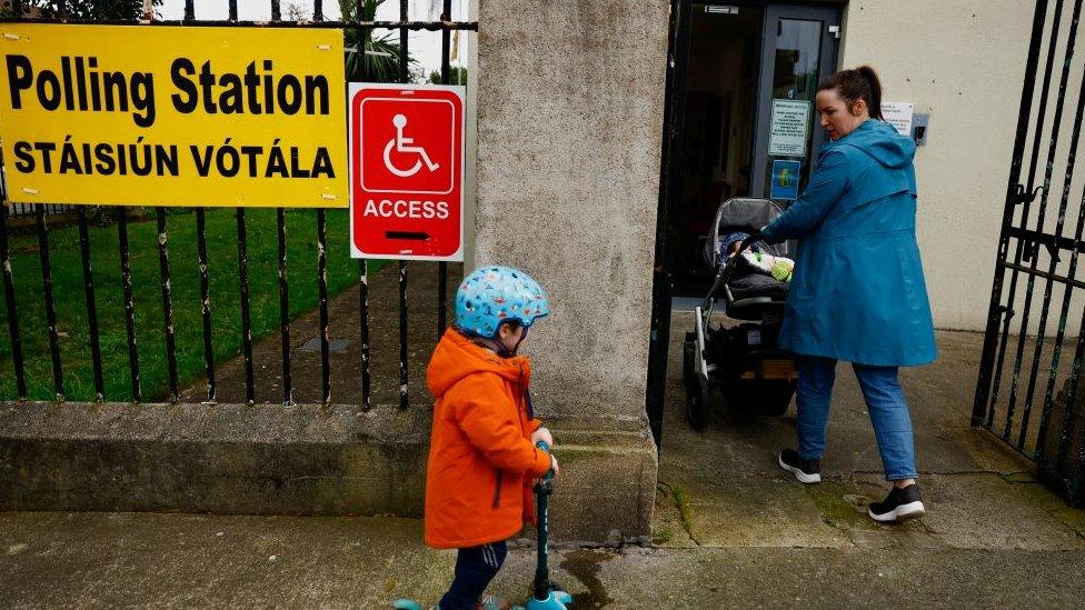 Mother pushing pram with child on scooter behind her entering polling station