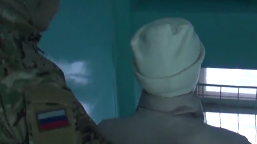 A screengrab of a video showing the woman wearing a white hat being escorted by a uniformed officer