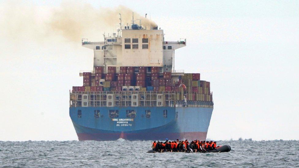 A small boat carrying migrants in front of a huge, industrial ship