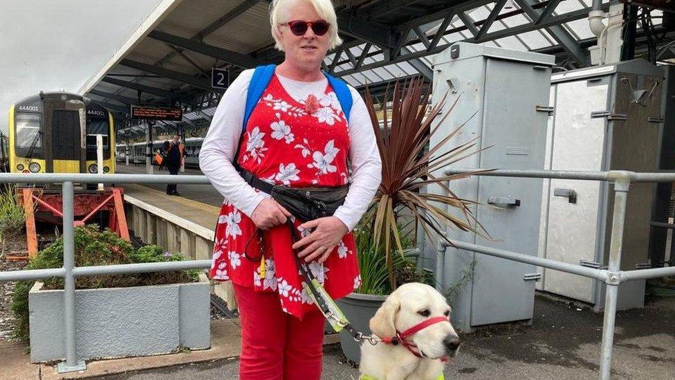 blind woman wearing red jeans standing with golden labrador guide dog at train station