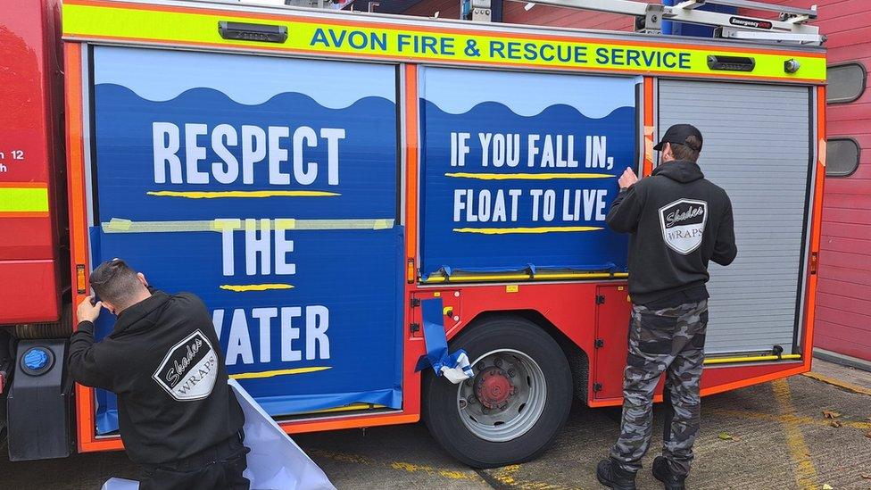 Two men putting the new wrap onto a fire engine. Their backs are to the camera. Both panels are royal blue with white capital letters which are underlined in yellow. The first panel reads 'Respect the water' and the second reads 'If you fall in, float to live'.