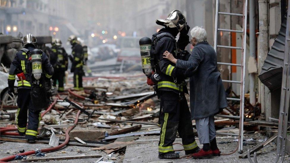 An elderly woman evacuated from a damaged building holds the arm of a fiefighter