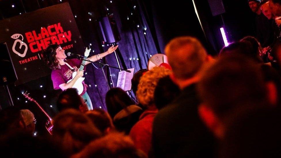 A woman playing a guitar in front of an audience at the Black Cherry Theatre