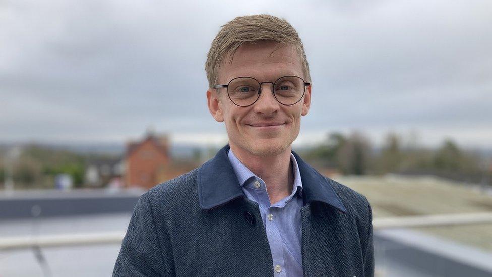 Jamie White smiles at the camera, wearing glasses and a dark blue coat