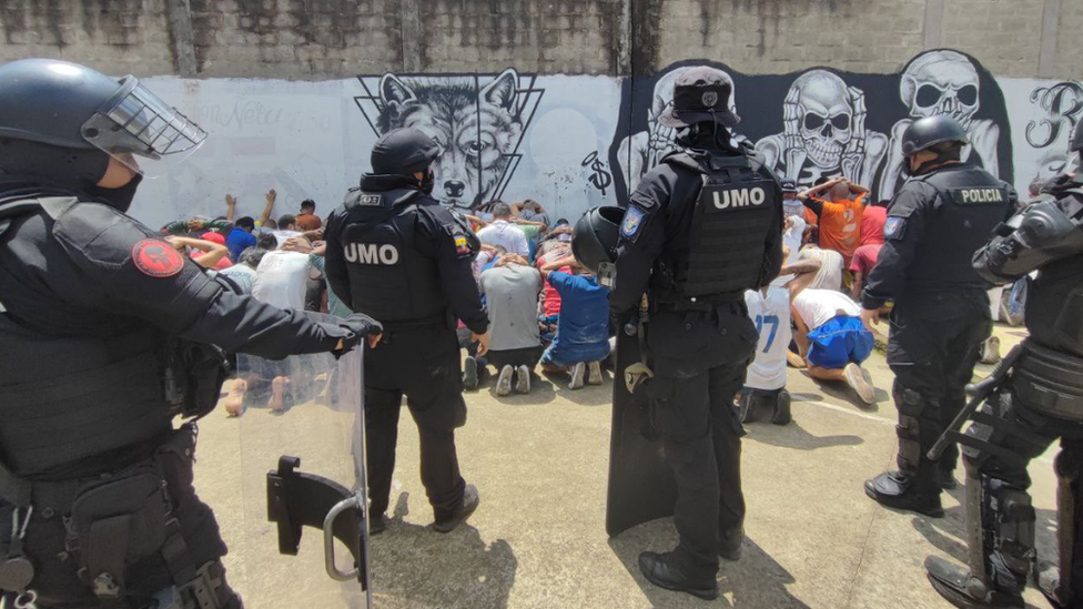 Heavily armed police guarding inmates on the ground with their hands behind their heads.