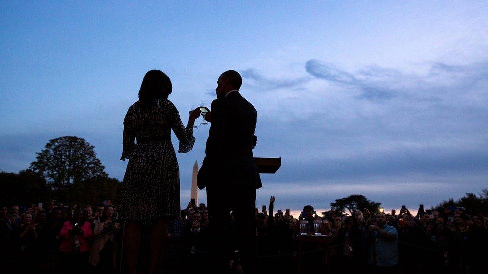 A silhouette at sunset shows Barack and Michelle Obama toast two champagne flutes before an assembled crowd, with the Washington Monument visible in the background