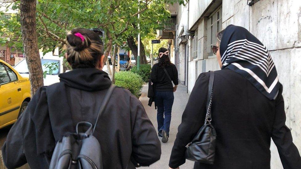 Two women walking down a street, facing away from the camera - one is wearing a headscarf and one is not