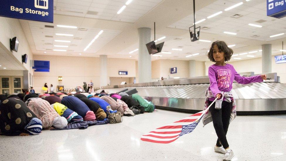 A young girl dances with an American flag in baggage claim while women pray behind her during a protest against the travel ban imposed by US President Donald Trump's travel ban at Dallas/Fort Worth International Airport