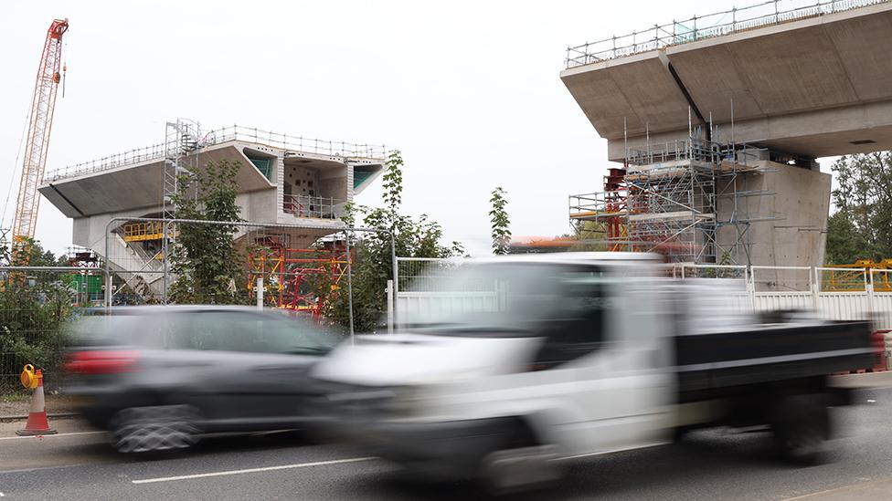 Construction takes place for the Colne Valley Viaduct section of the HS2 high speed rail project in in Denham, Britain, 28 September 2023. A bridge being built in background with a car and a truck driving in the foreground.