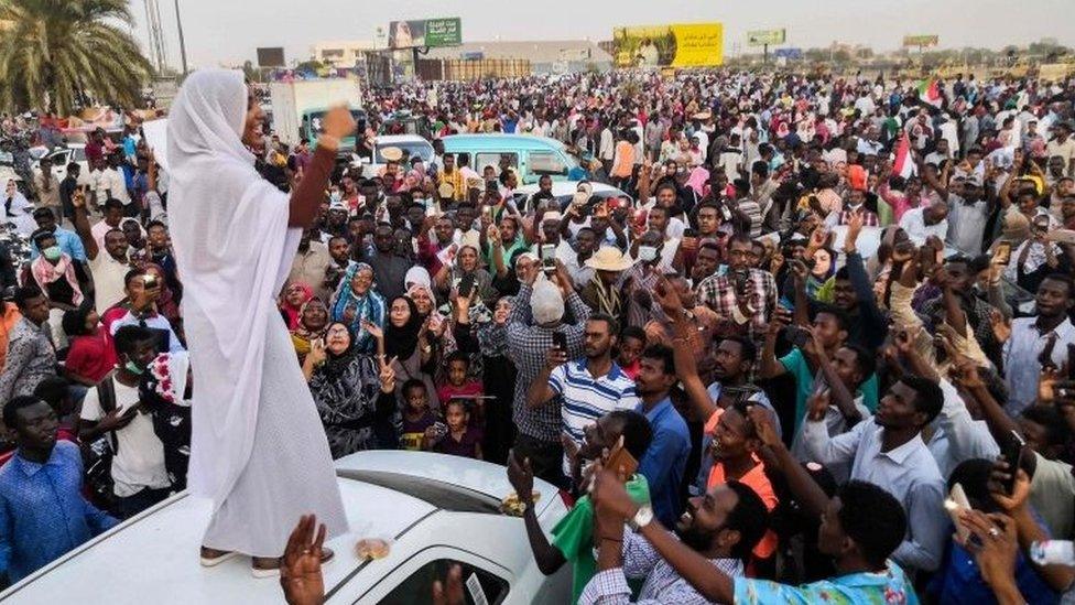 Protesters in Khartoum hold up their phones to film fellow protester Alla Salah who stands on a car dressed in a white headscarf and skirt