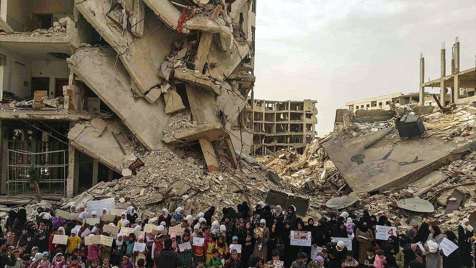 Residents of Darayya hold signs amid destroyed buildings, calling for help from the international community