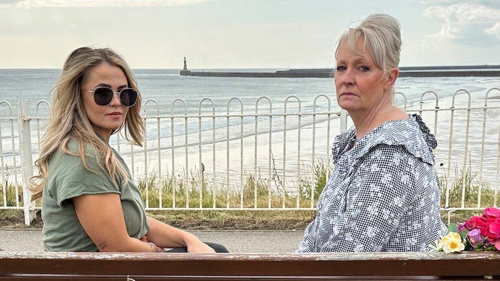 Two women sit on a bench overlooking the sea