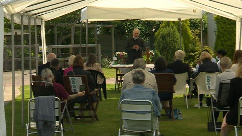 A vicar leads people gathered at a memorial service in a tent in the grounds of the care home.