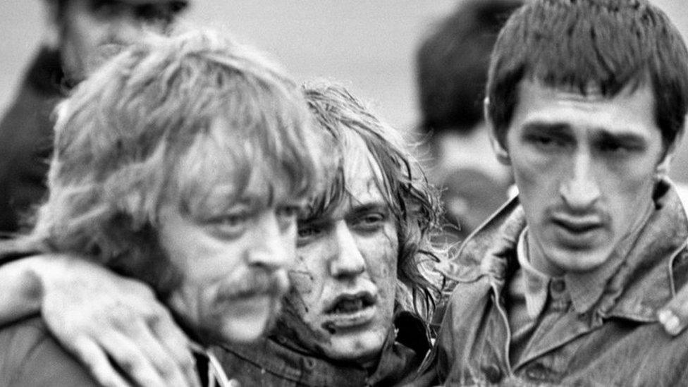 A man with a bloodied face puts his arms around the shoulders of two men helping him