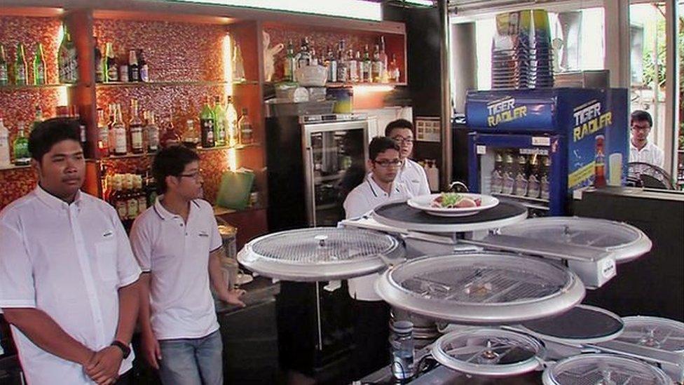 Waiters stand by drones holding food.