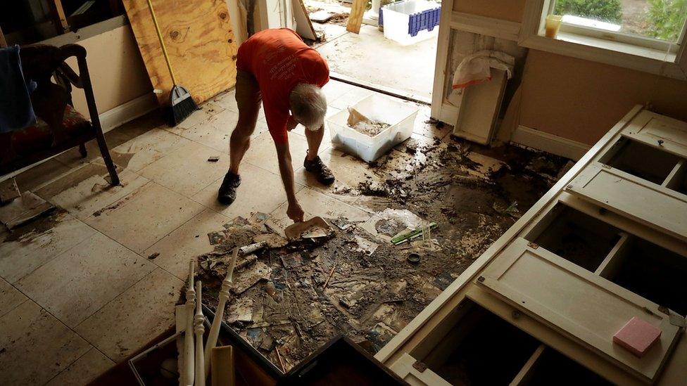 An elderly man bends down to clear debris and mud from his home