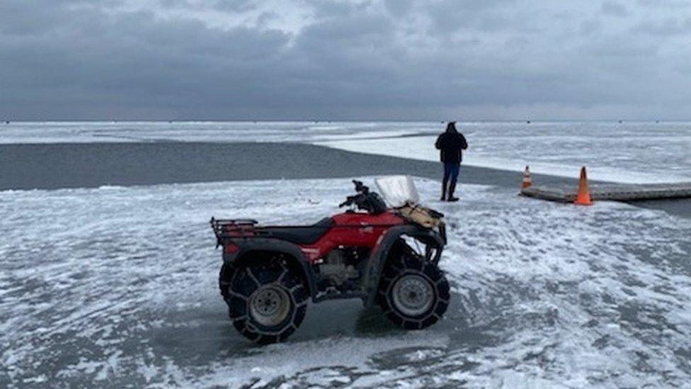 Photo of a rescue vehicle on the ice next to a narrow part of open water with a portable bridge placed atop it so people can cross.