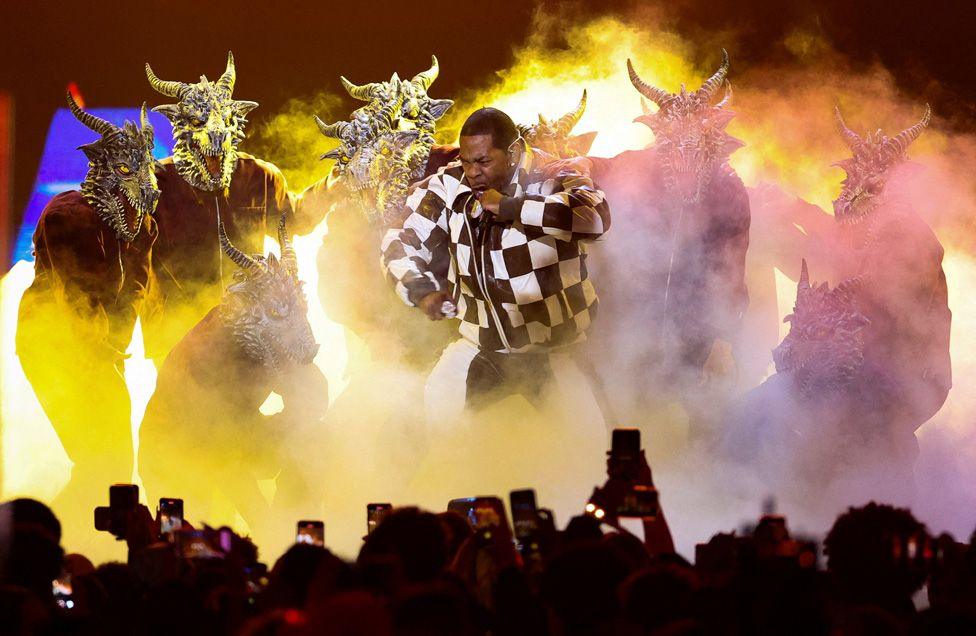 Busta Rhymes performing in the middle of dancers wearing horned animal masks, and dry ice and yellow lights