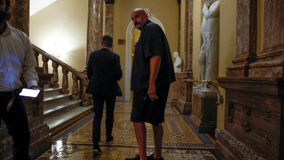 Fetterman in dark short-sleeved shirt, shorts and sneakers in a yellow hallway next to man in suit.