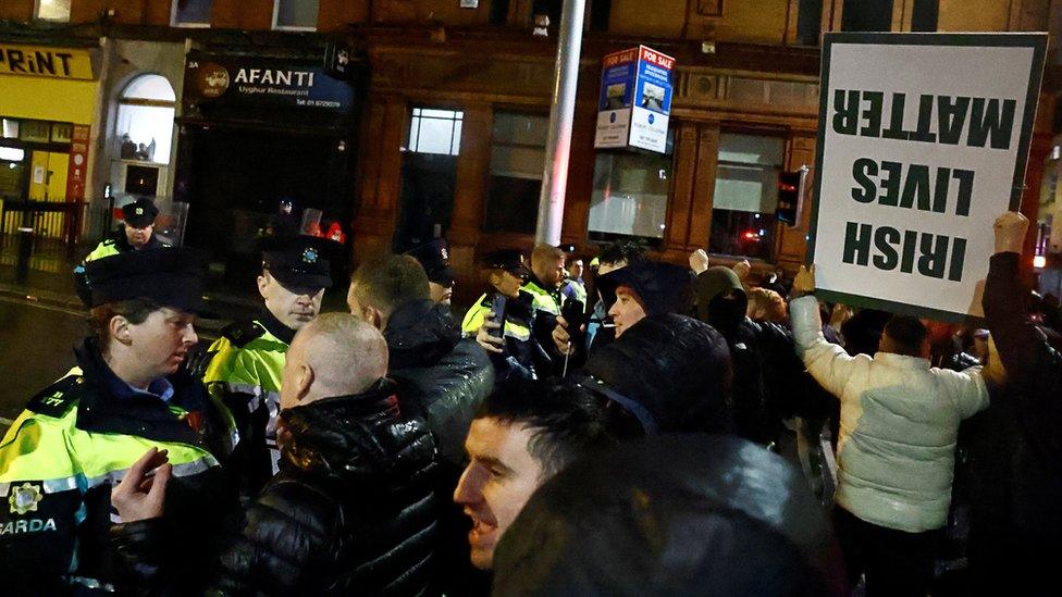 Police officers form a line as a crowd of people gather in Dublin city centre, with one man holding a sign that reads: IRISH LIVES MATTER