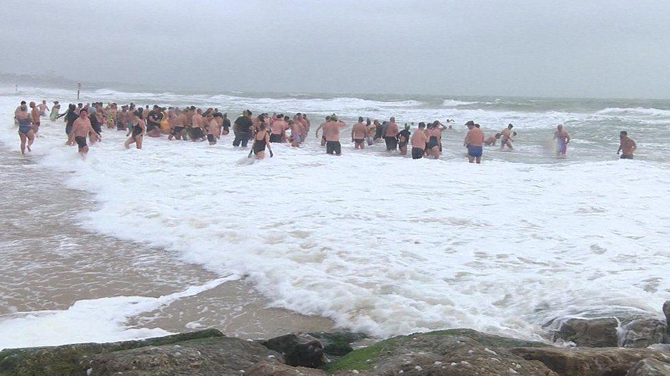 Rocks along a frothing sea with dozens of people wading out into the sea.