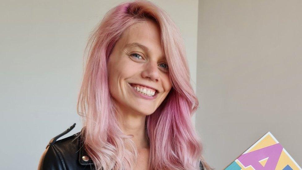 Leanne has a big smile and she's looking straight at the camera. She's got long pink wavy hair and is wearing a black leather jacket and is holding her book she's written about ADHD, but it's slightly cropped out of the shot.