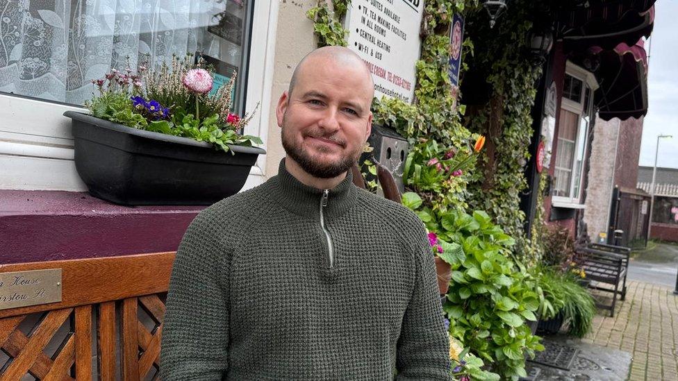 Andrew Cregan, a shaven-headed man with a small brown beard, sits in front of flowers