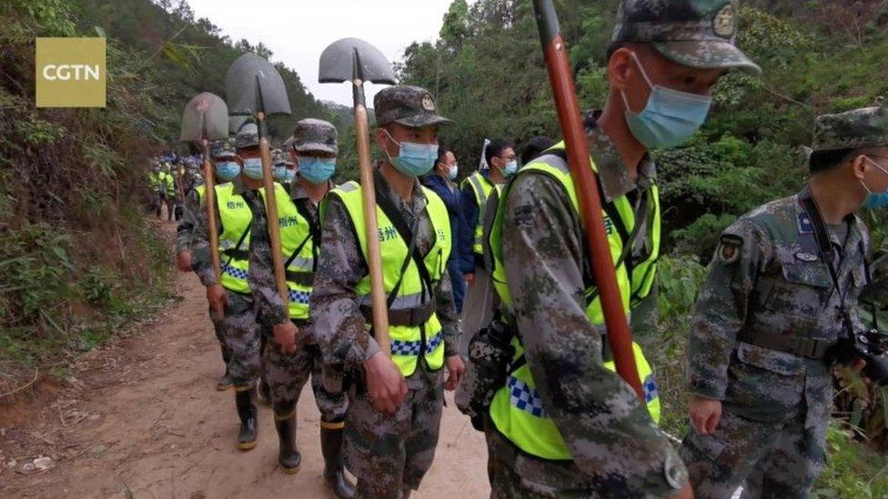 Soldiers carrying shovels walk along a pathway to the forested mountain crash site