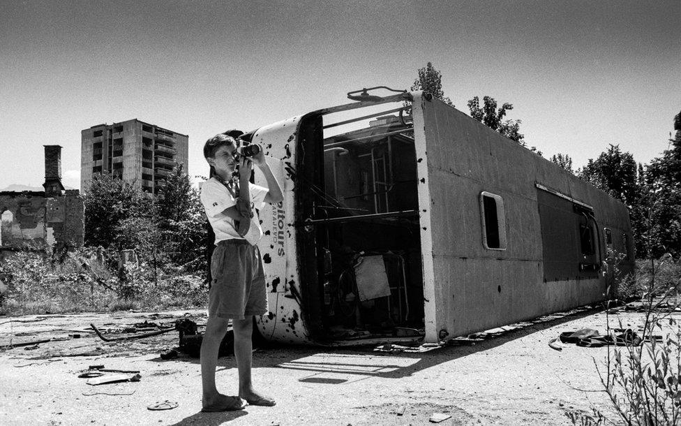 A child holds a camera next to a destroyed bus