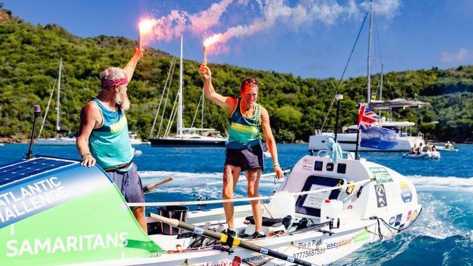 Simon and Nina Crouchman stand up in boat holding flares to celebrate arriving in Antigua after completing Atlantic row challenge