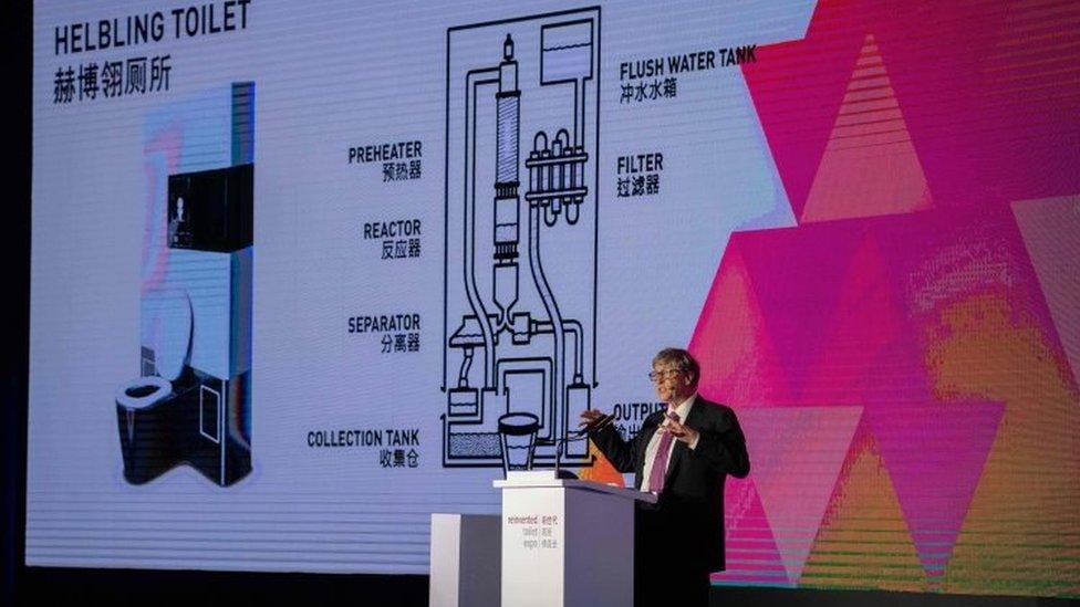 Bill Gates stands in front of a screen with a model of a 'Helbling' toilet at event