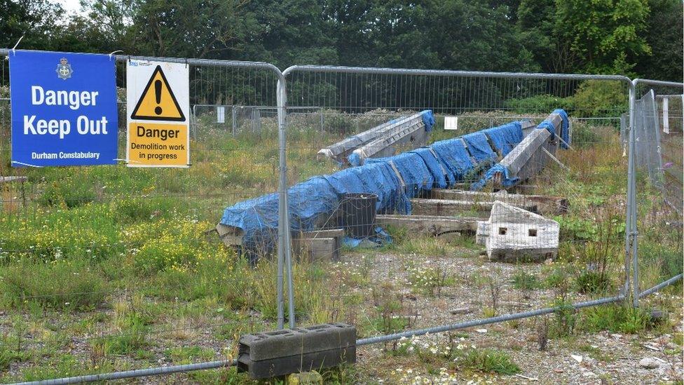 Concrete structures lie on the ground beneath blue covers surrounded by metal fencing