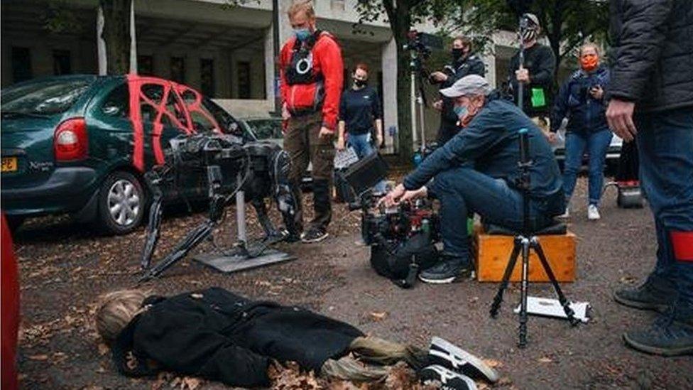 An actor lies on the floor in a chaotic street scene, with the camera operator and crew wearing masks