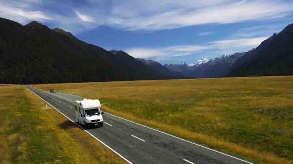 Still from the promotional video showing a white camper van driving on the right side of the road, with mountains in the background