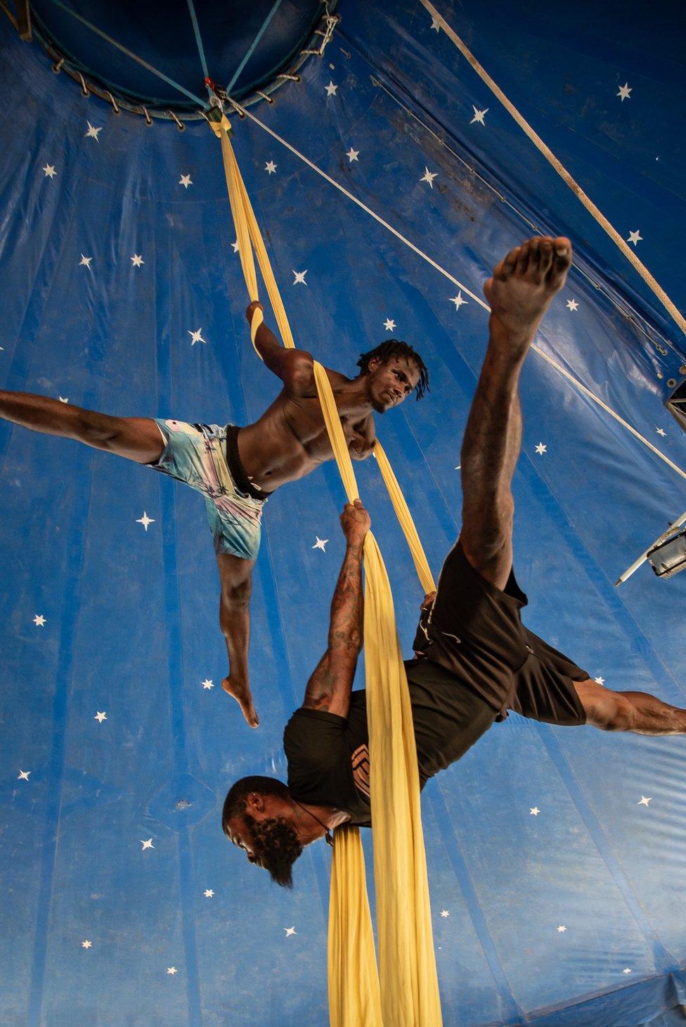 Two acrobats suspend themselves mid-air with yellow aerial silks.