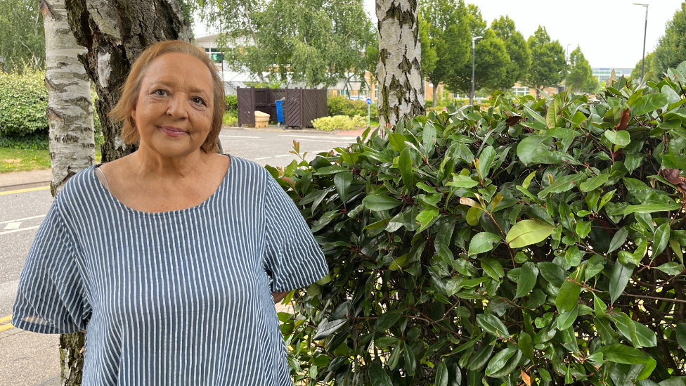 Linda Huskisson is stood outdoors and to the left side of a bush with lots of green leaves on it. She has light brown hair that reaches the top of her shoulders and is smiling at the camera. She wears a vertically striped blue and white dress.